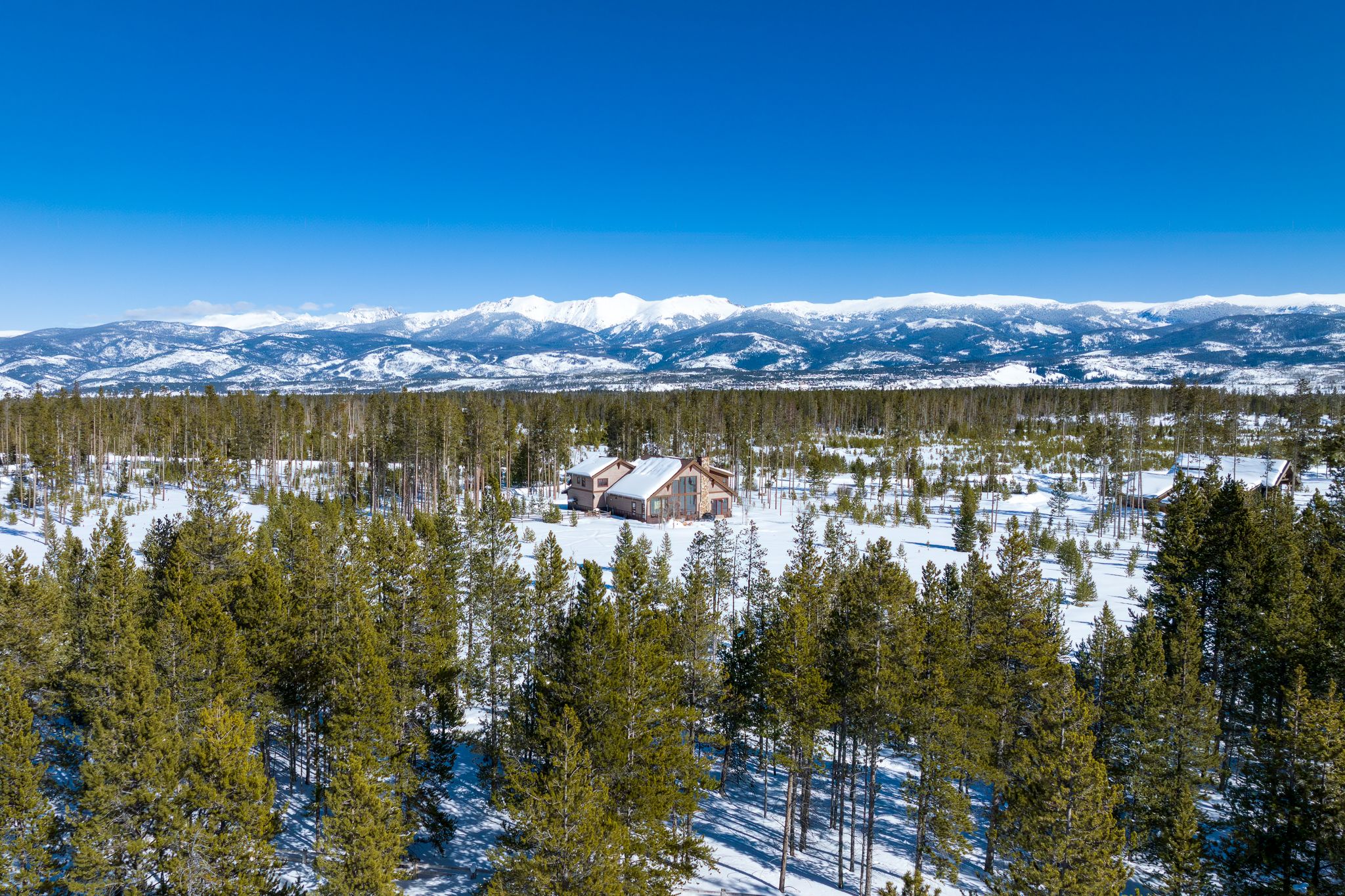 A snowy mountain landscape with a large cabin nestled among pine trees, surrounded by vast forest and distant snow-capped peaks under a clear blue sky.
