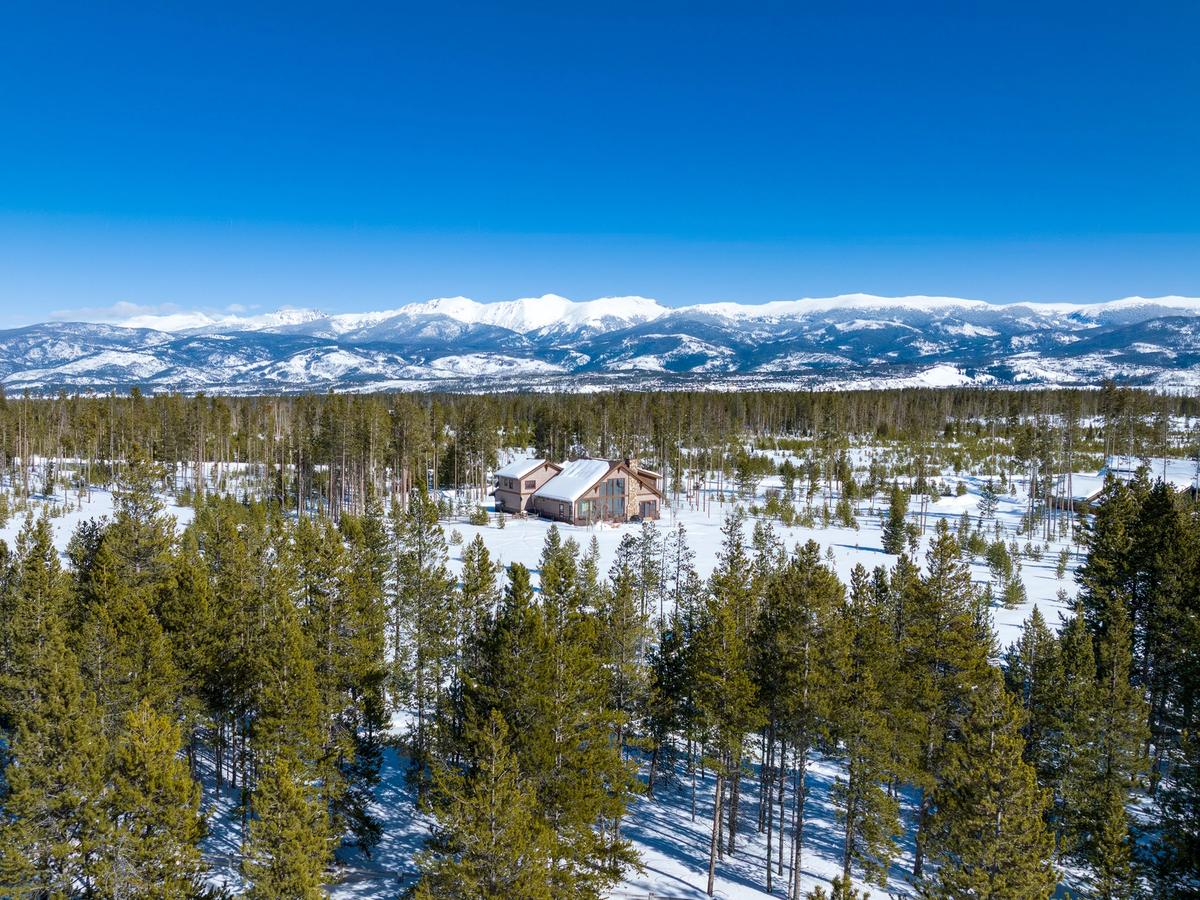 A snowy mountain landscape with a large cabin nestled among pine trees, surrounded by vast forest and distant snow-capped peaks under a clear blue sky.