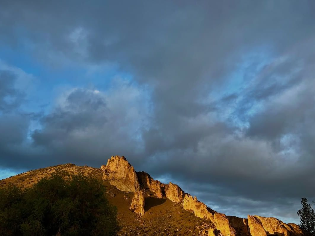 Golden cliffs of Smith Rock illuminated by sunlight against a dramatic cloudy sky, showcasing Oregon’s high desert beauty.