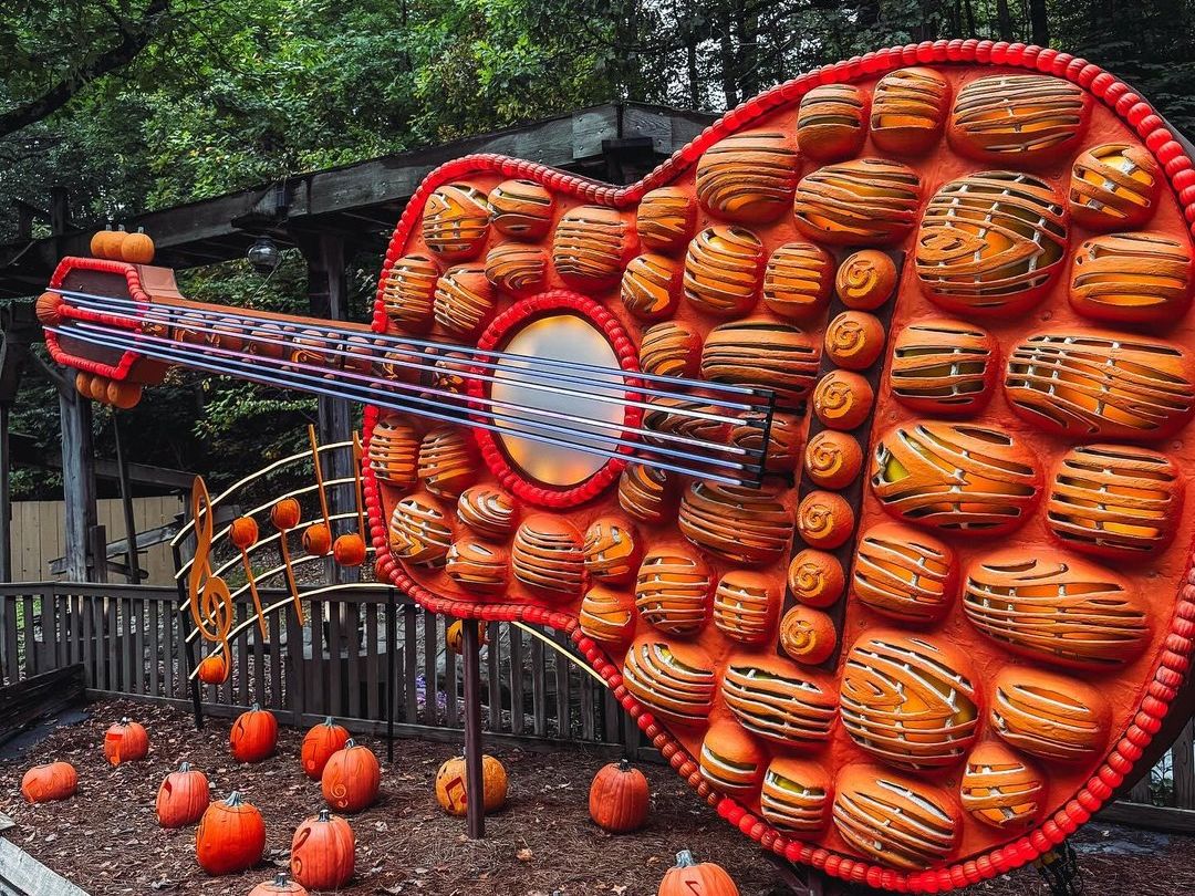 A giant guitar made of orange pumpkins stands at Dollywood surrounded by smaller pumpkins and trees. The festive fall display celebrates the park’s love for music and autumn fun.