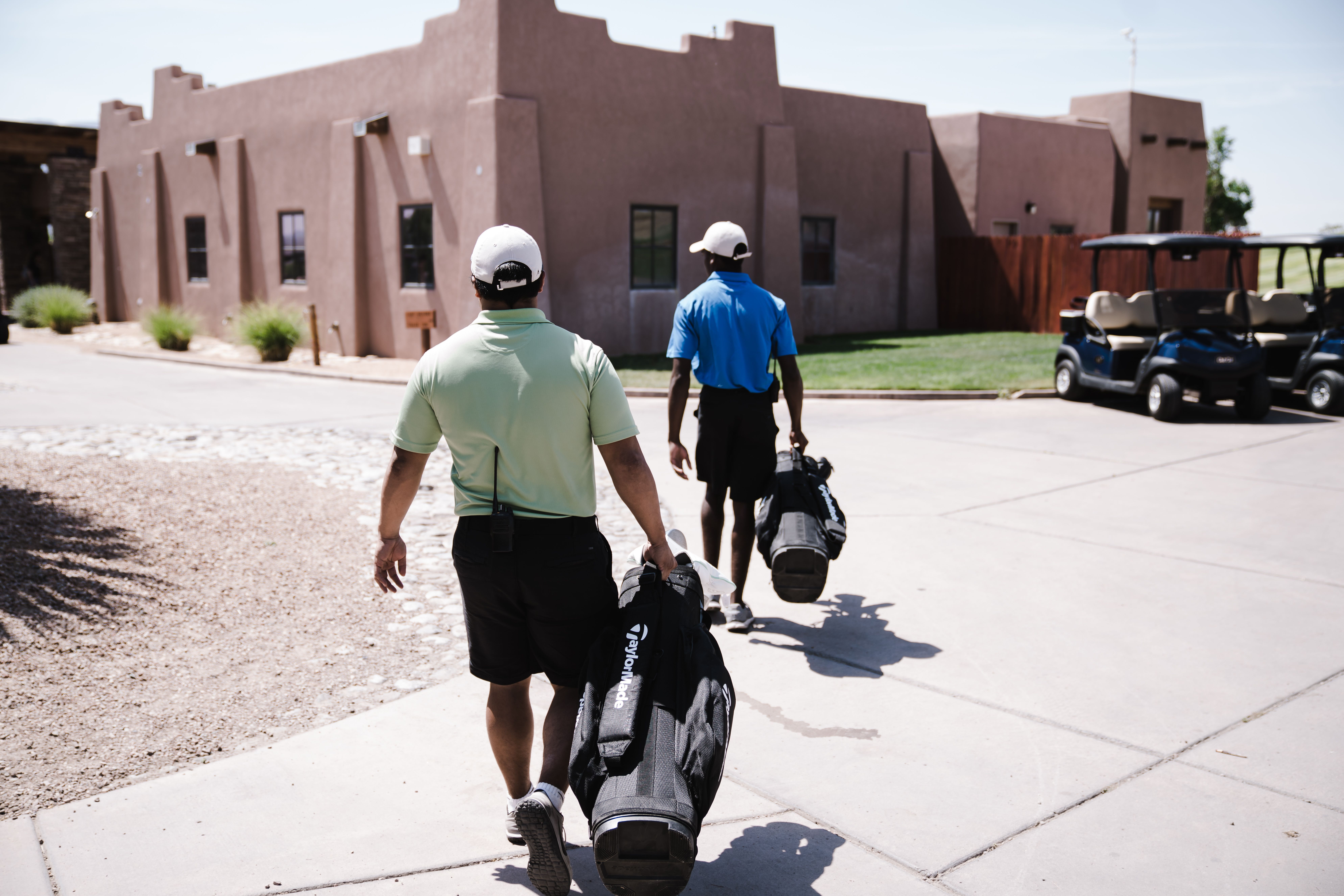 Two golfers carrying TaylorMade and stand bags walk toward a southwestern-style adobe clubhouse on a sunny day, heading out for a round at a resort golf course. 