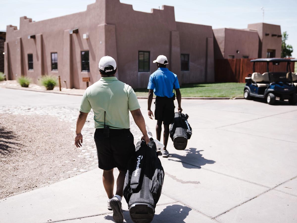 Two golfers carrying TaylorMade and stand bags walk toward a southwestern-style adobe clubhouse on a sunny day, heading out for a round at a resort golf course.