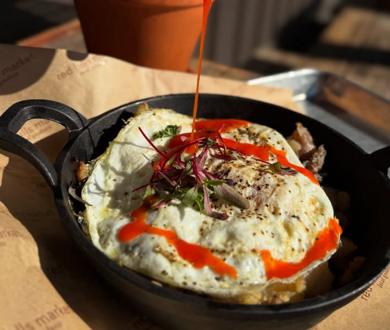 A skillet breakfast topped with two fried eggs, microgreens, and a drizzle of bright red hot sauce being poured on top.