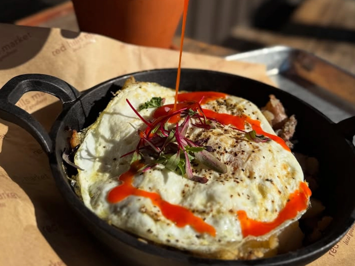 A skillet breakfast topped with two fried eggs, microgreens, and a drizzle of bright red hot sauce being poured on top.