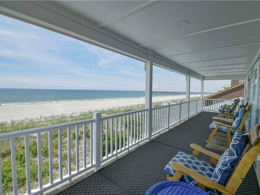 A wide oceanfront porch at Holden Beach with wooden rocking chairs lined up, offering peaceful views of the sandy shore and blue waves. Perfect spot to relax and enjoy the breeze by the water.