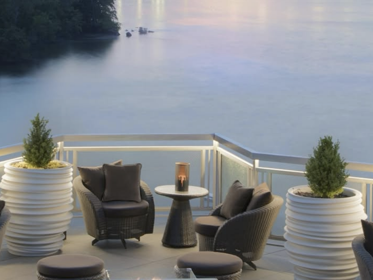 Cozy rooftop lounge with modern wicker chairs, potted greenery, and a candlelit table overlooking a calm river at dusk.