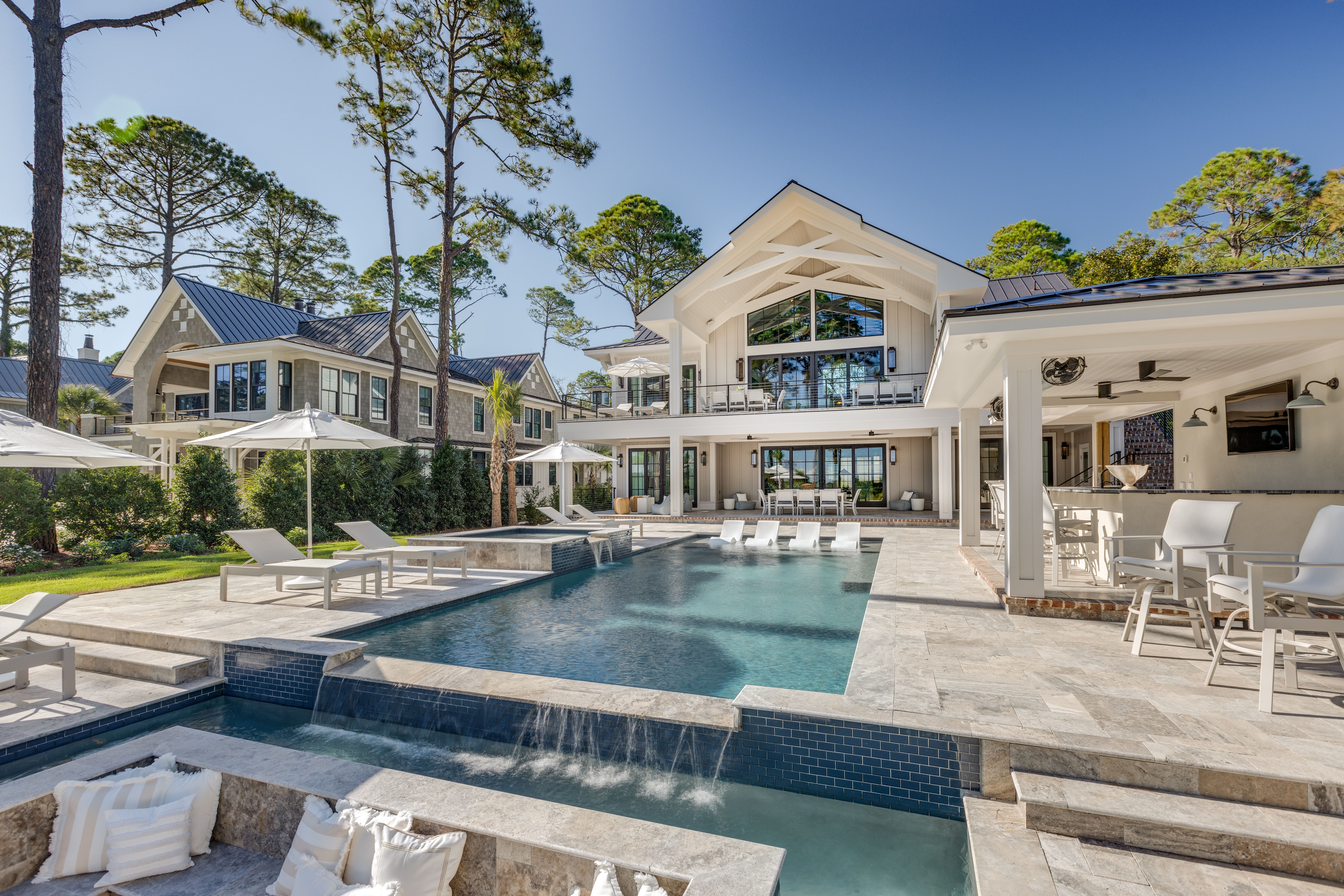 A luxurious coastal estate features an expansive stone patio with a multi-level pool, in-water loungers, a covered outdoor bar, and modern lounge areas, framed by tall pine trees and elegant architecture.