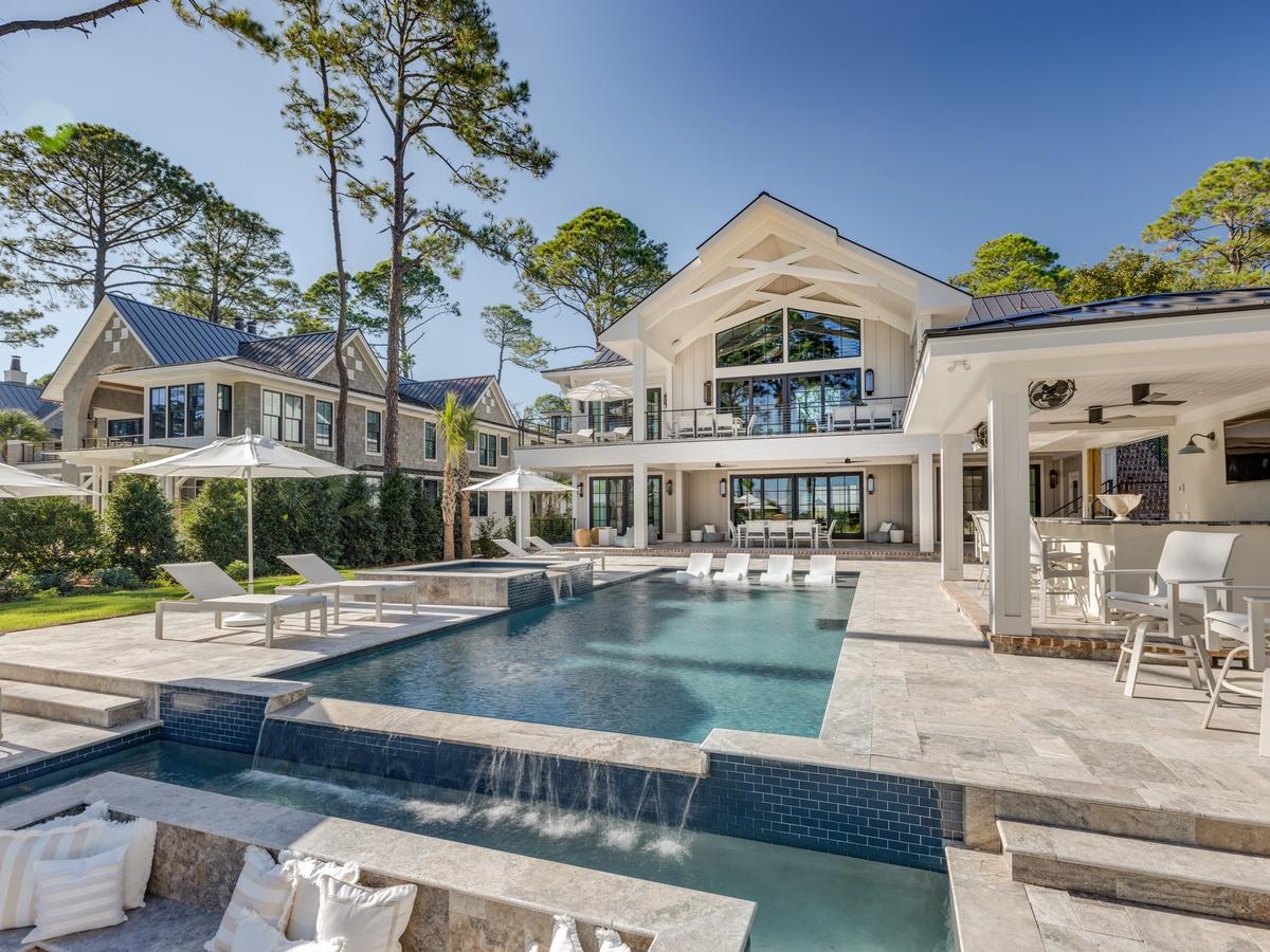 A luxurious coastal estate features an expansive stone patio with a multi-level pool, in-water loungers, a covered outdoor bar, and modern lounge areas, framed by tall pine trees and elegant architecture.