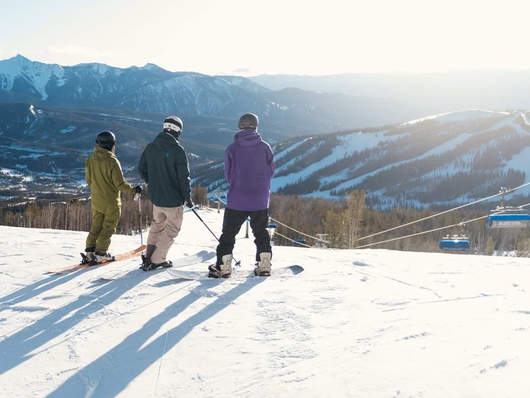 Three snowboarders stand at the top of a snowy slope, taking in the mountain view before heading down. Big Sky’s winter magic is in full display with wide trails and fresh powder for every level of rider.
