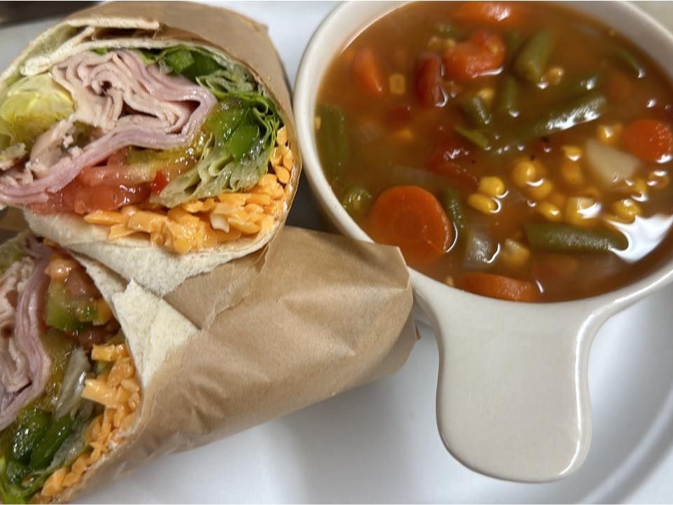A colorful veggie-packed wrap with ham, cheddar cheese, and fresh lettuce, served next to a bowl of hearty vegetable soup. A comforting lunch combo full of color and crunch.