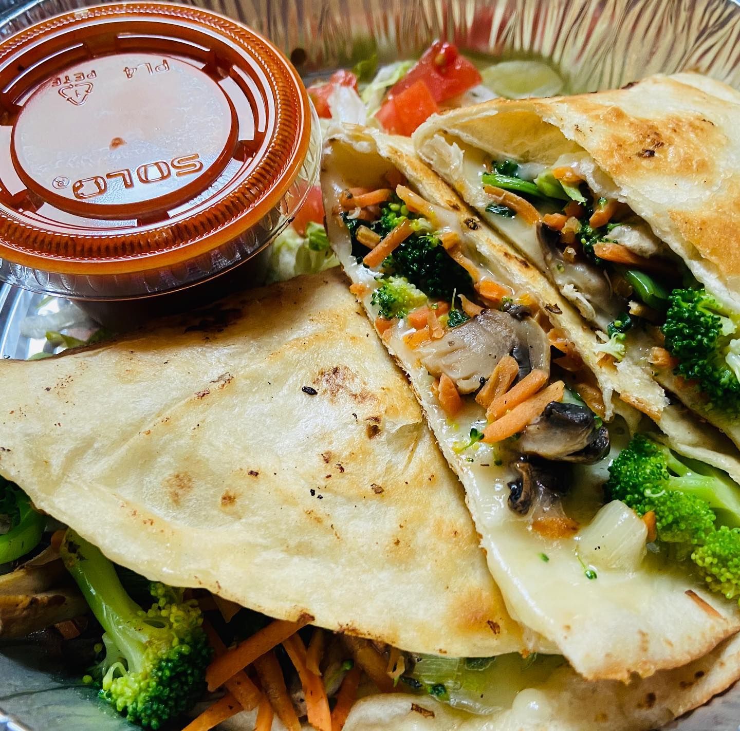 A golden-griddled quesadilla is cut open to reveal a filling of melted cheese, broccoli, mushrooms, and shredded carrots, served in a foil tray. A cup of red salsa and a side of fresh lettuce and diced tomato round out the plate.