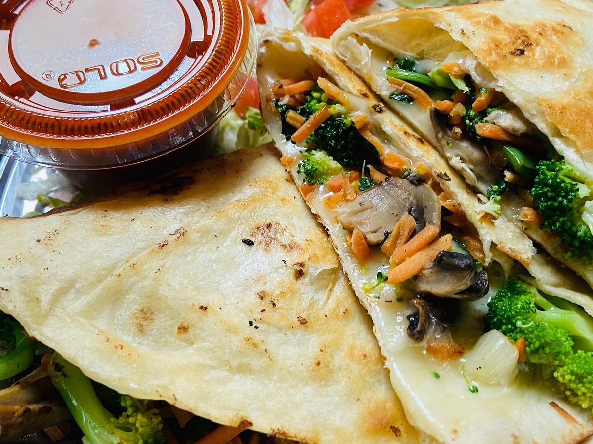 A golden-griddled quesadilla is cut open to reveal a filling of melted cheese, broccoli, mushrooms, and shredded carrots, served in a foil tray. A cup of red salsa and a side of fresh lettuce and diced tomato round out the plate.