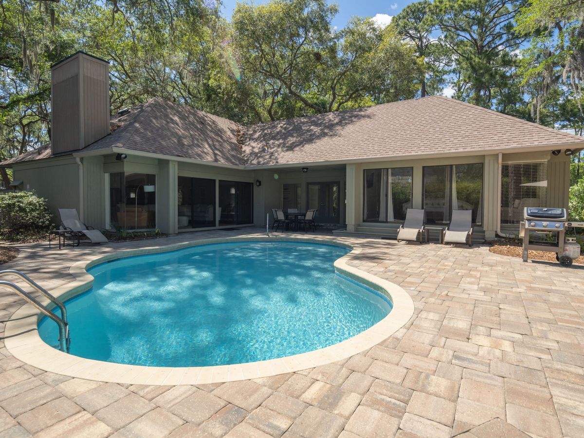 A super stylish, fully renovated coastal home features a bright open-plan living area with modern white sofas, crisp kitchen finishes, and a private pool with custom evening lightscaping, set against serene lagoon and golf views.