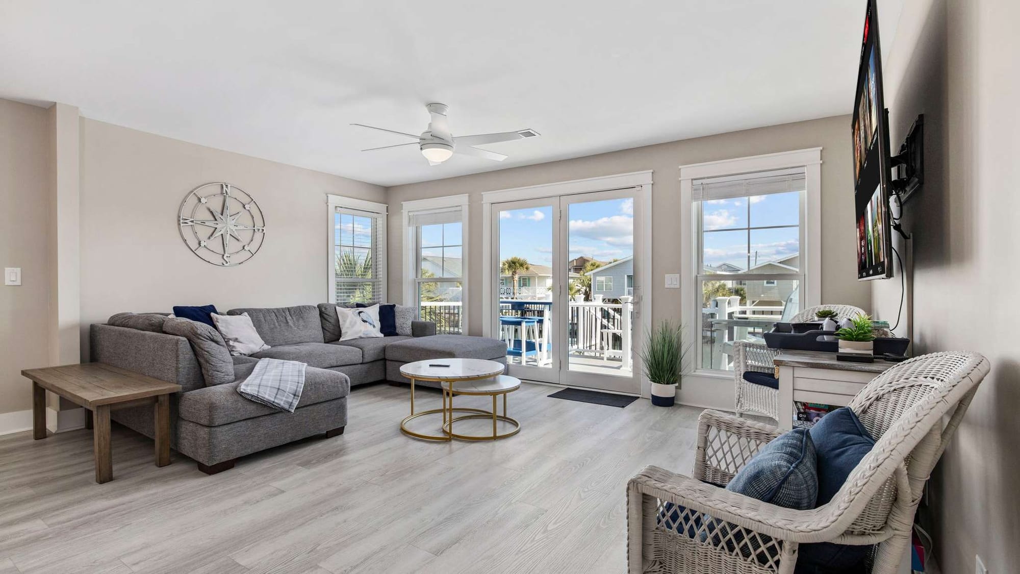 Bright coastal living room with large windows and balcony access brings in natural light and relaxing neighborhood views. The comfortable sectional seating makes this space perfect for family gatherings during a beach vacation.