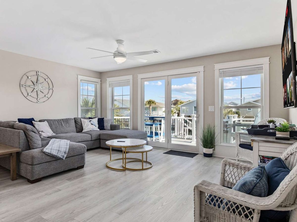 Bright coastal living room with large windows and balcony access brings in natural light and relaxing neighborhood views. The comfortable sectional seating makes this space perfect for family gatherings during a beach vacation.