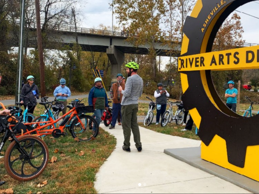 A group of cyclists wearing helmets gather around a guide near the large yellow and black “River Arts District” sign in Asheville, North Carolina.