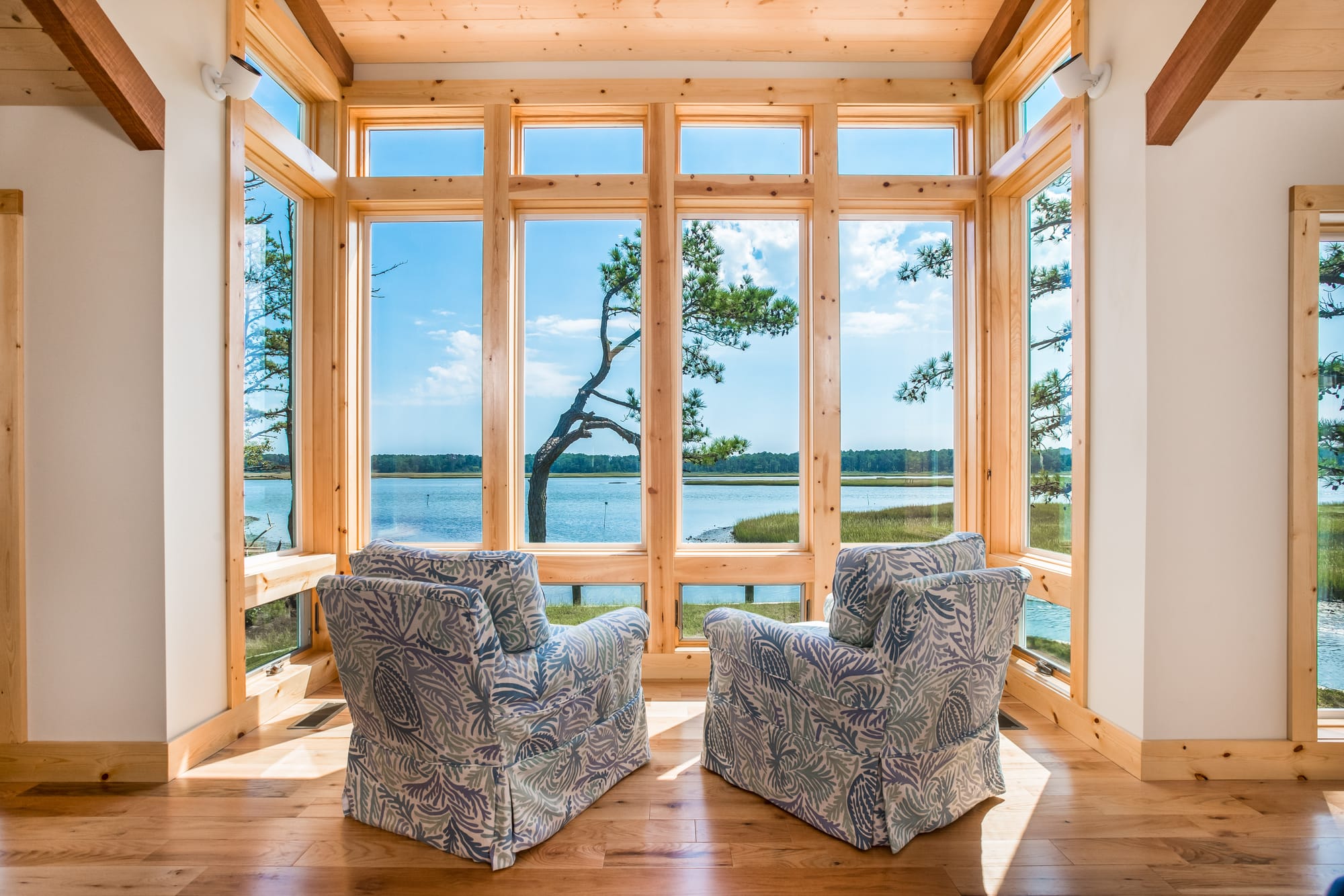Two patterned armchairs overlook a peaceful waterfront through tall wood-framed windows in Chincoteague, Virginia. Sunlight fills the cozy room, creating the perfect spot to unwind and enjoy the view.