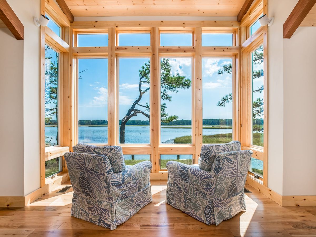 Two patterned armchairs overlook a peaceful waterfront through tall wood-framed windows in Chincoteague, Virginia. Sunlight fills the cozy room, creating the perfect spot to unwind and enjoy the view.