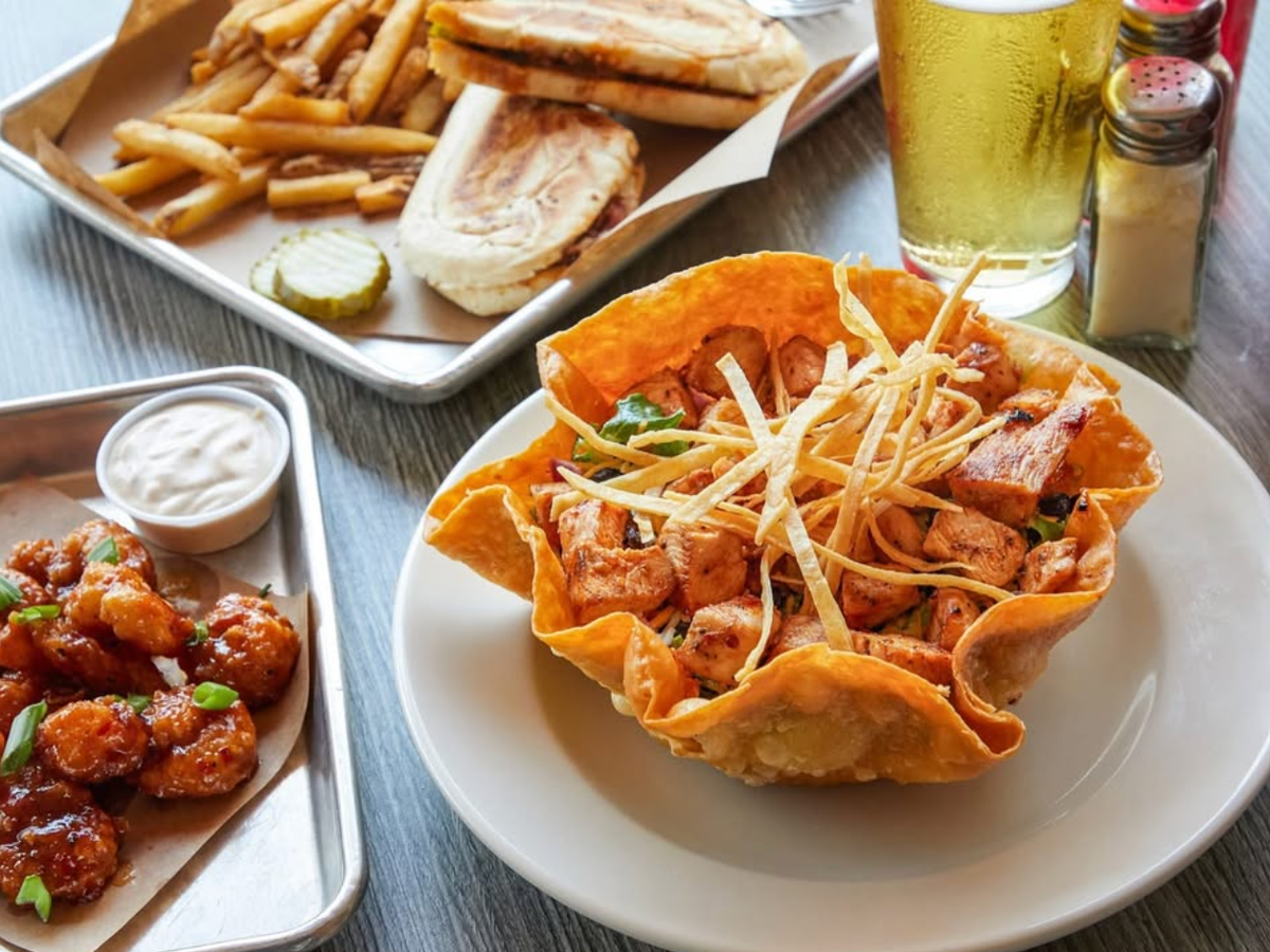 A crispy taco bowl filled with grilled chicken and topped with tortilla strips, served alongside a tray of saucy shrimp with dipping sauce, a Cuban sandwich with fries and pickles, and a cold glass of beer.
