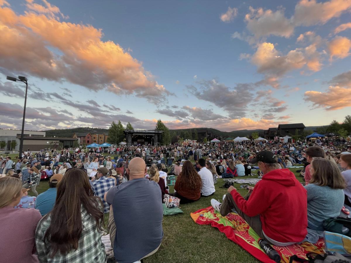 A large crowd gathers on a grassy field to enjoy a live concert under a colorful sunset sky. The relaxed, festival vibe and mountain backdrop create a perfect summer evening scene.