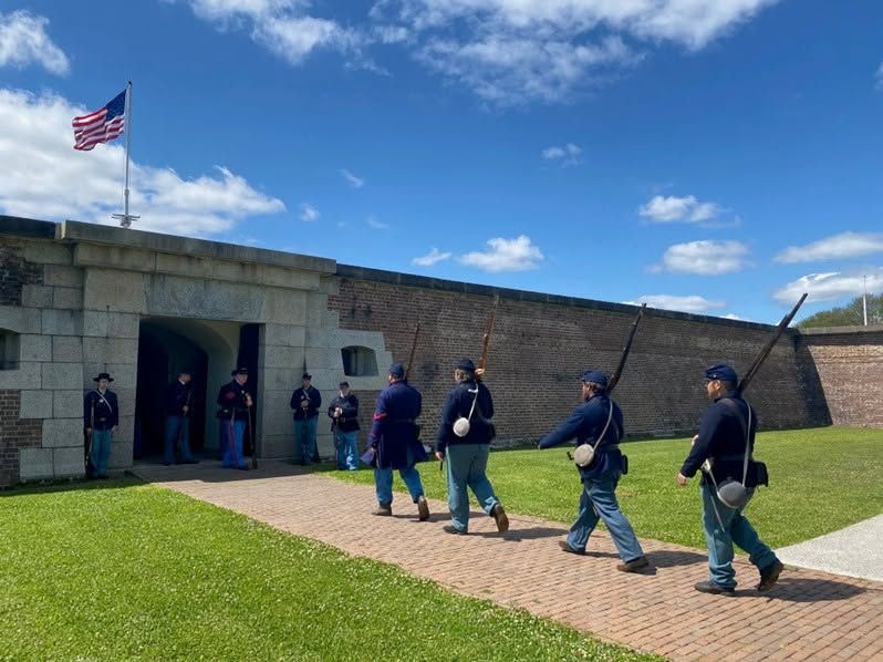 Civil War reenactors in Union Army uniforms march in formation across the brick courtyard of Fort Sumter National Monument in Charleston Harbor, with the American flag flying above the historic fortification walls on a sunny spring day.