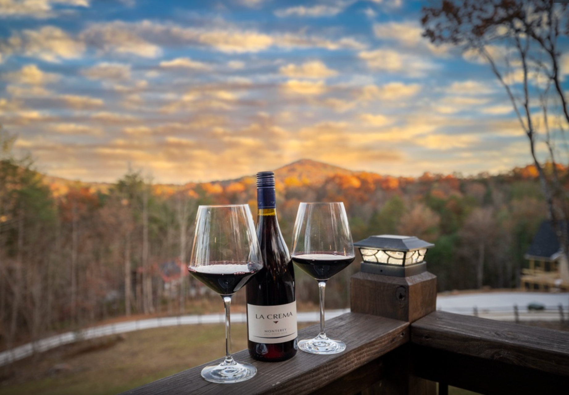 Two glasses of red wine and a bottle sit on a wooden deck overlooking a mountain landscape at sunset with autumn foliage.