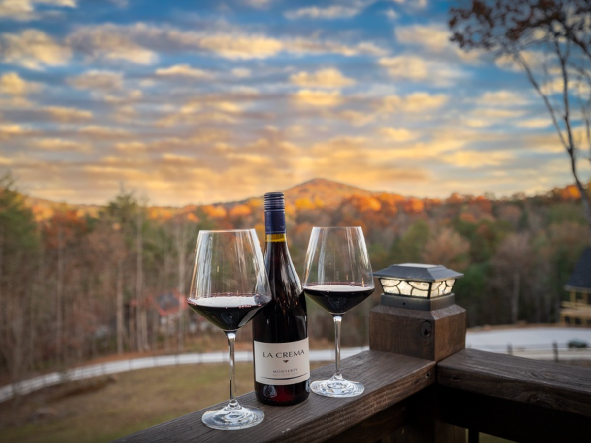 Two glasses of red wine and a bottle sit on a wooden deck overlooking a mountain landscape at sunset with autumn foliage.