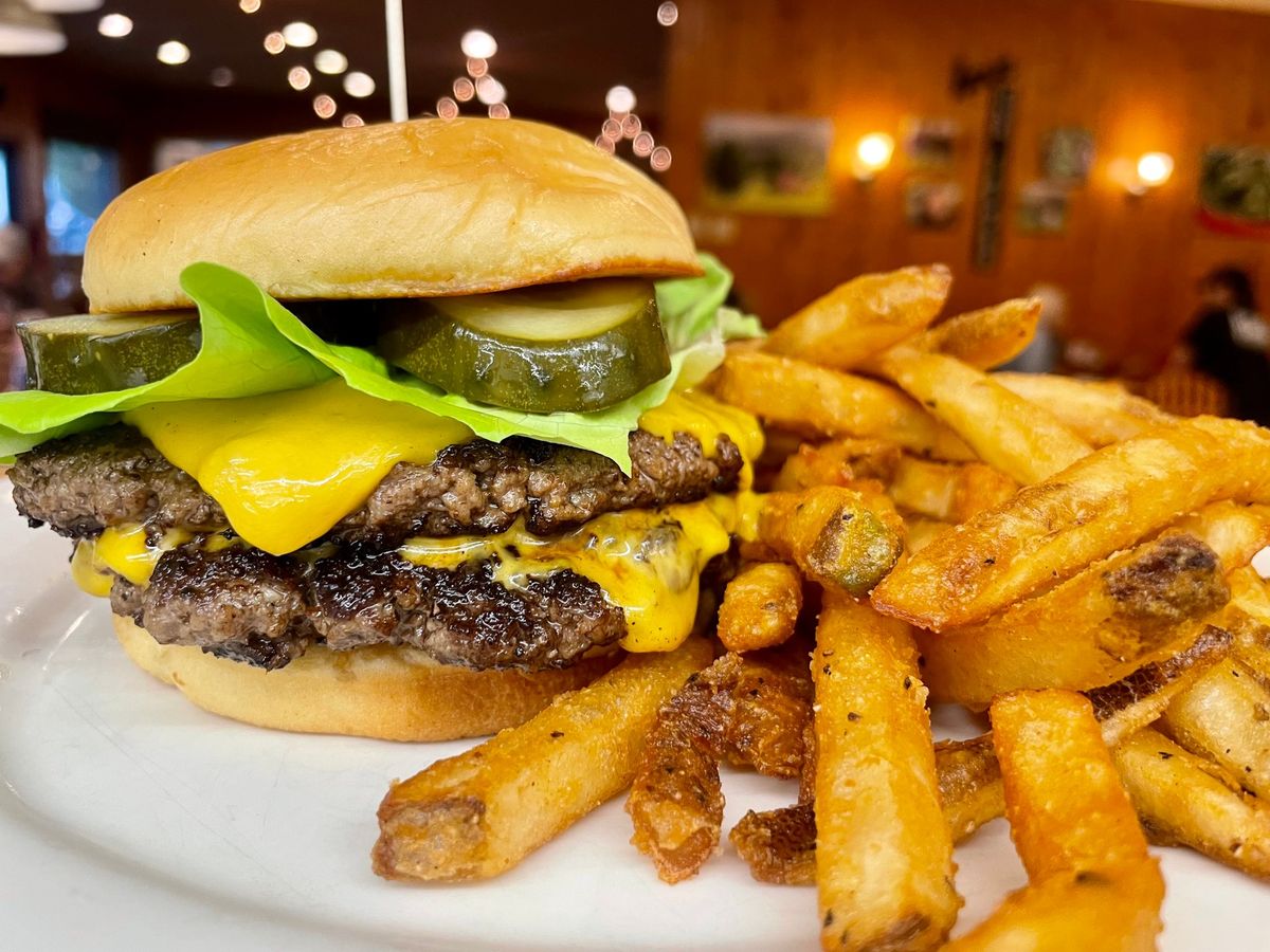 A juicy double cheeseburger topped with pickles and crisp lettuce sits beside golden fries. The cozy, rustic restaurant atmosphere adds to the charm of this hearty Southern-style meal.