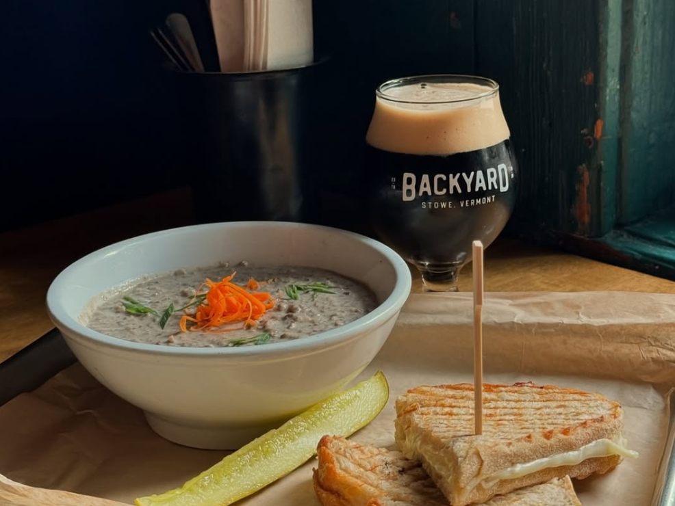 A cozy meal of creamy soup, a grilled cheese sandwich, and a pickle spear is served on a tray beside a dark stout from Backyard in Stowe, Vermont