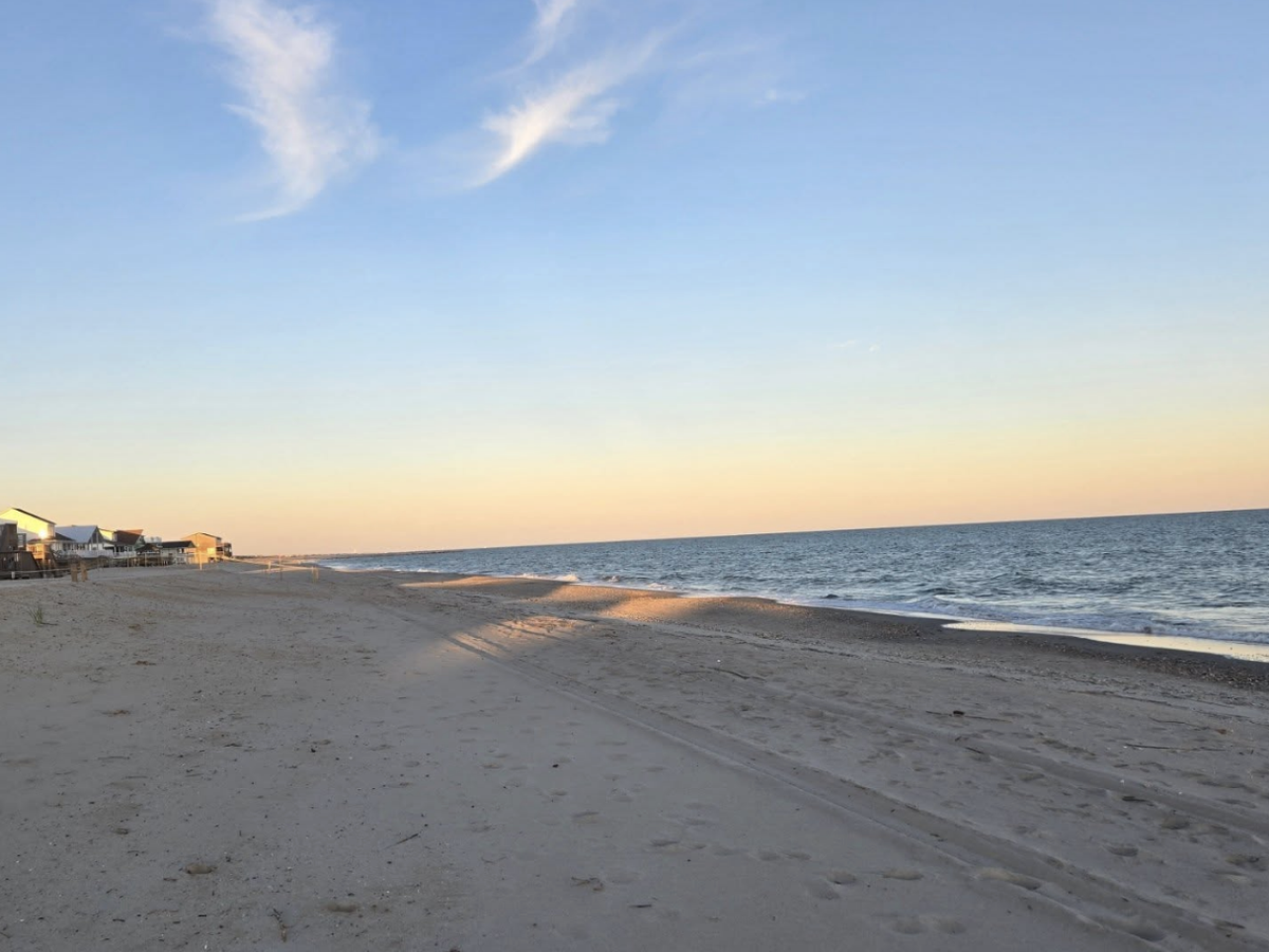A peaceful beach at sunset with soft golden light reflecting on the sand and water, and beach houses lining the shoreline in the distance.