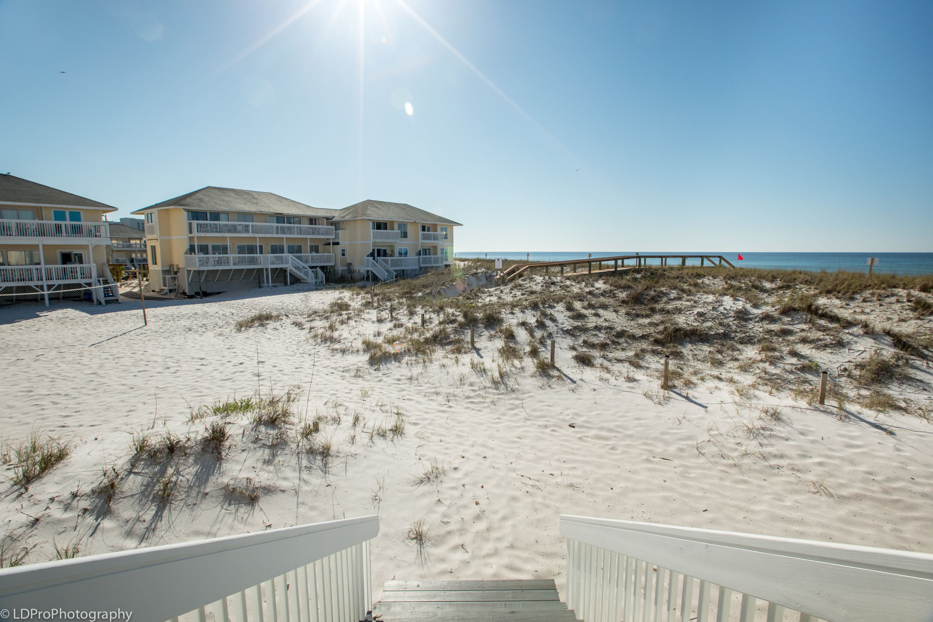 View from a wooden staircase leading down to a sunlit, sandy beach with grassy dunes, yellow beachfront cottages, and a wooden boardwalk extending toward the ocean.