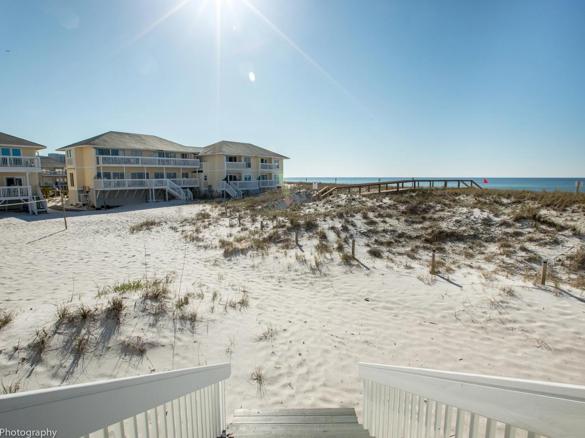 View from a wooden staircase leading down to a sunlit, sandy beach with grassy dunes, yellow beachfront cottages, and a wooden boardwalk extending toward the ocean.