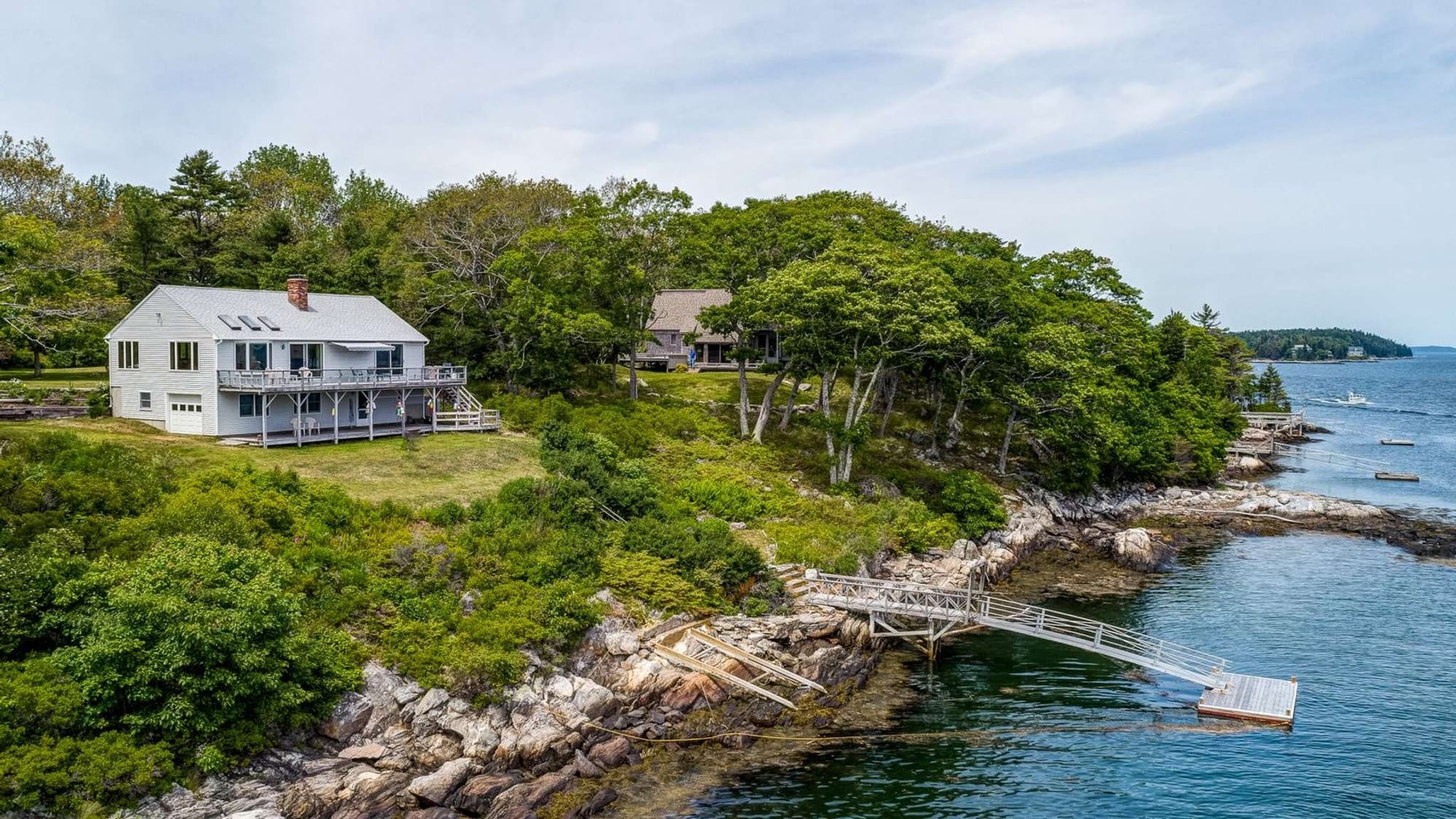 A scenic rocky coastline with pine trees and waterfront homes overlooking the ocean. Gentle waves roll along the shoreline while boats drift in the distance. Boothbay is known for its peaceful harbor views and classic coastal New England scenery.
