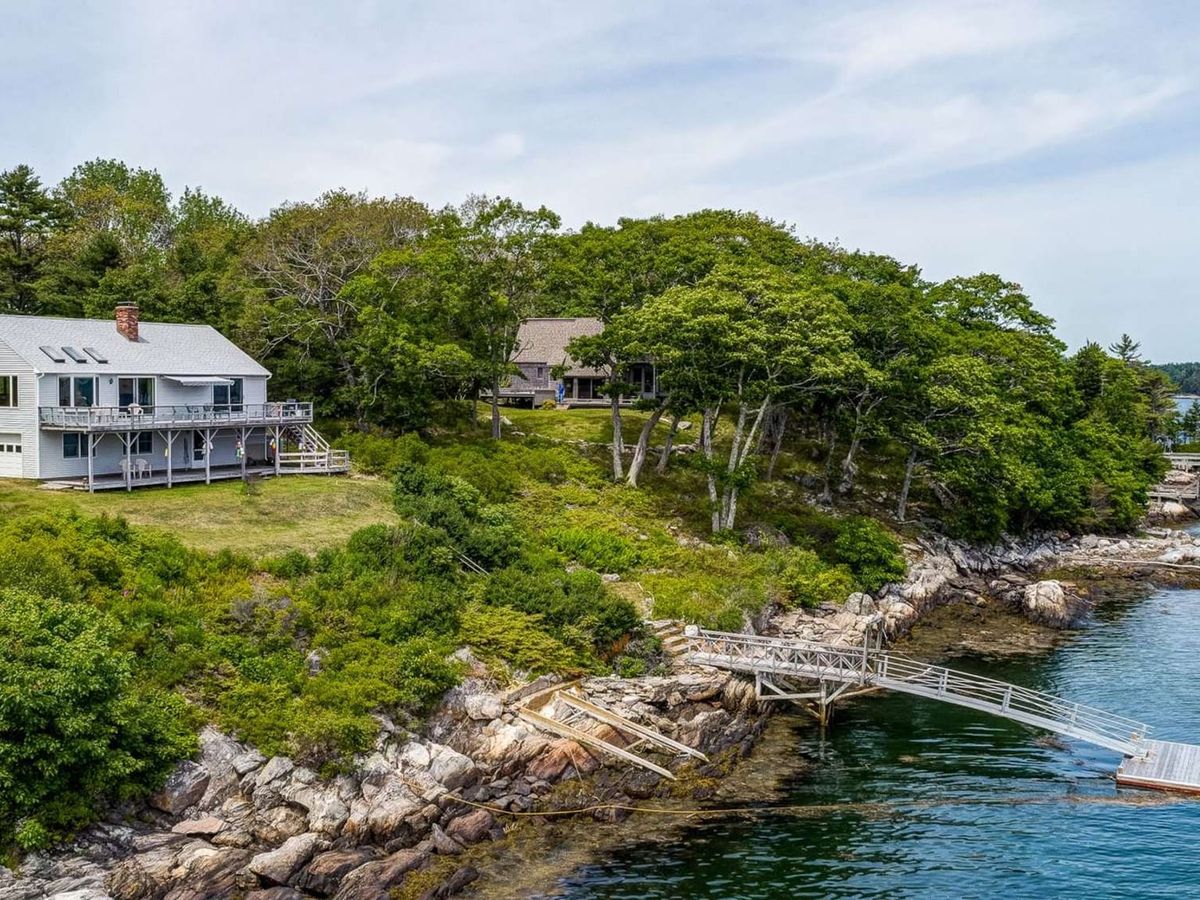 A coastal waterfront home in Maine sits on a rocky shoreline with a private dock leading down to the clear blue water. Surrounded by trees and ocean views, this scenic property offers peaceful seaside living and easy access for boating and relaxing by the shore.