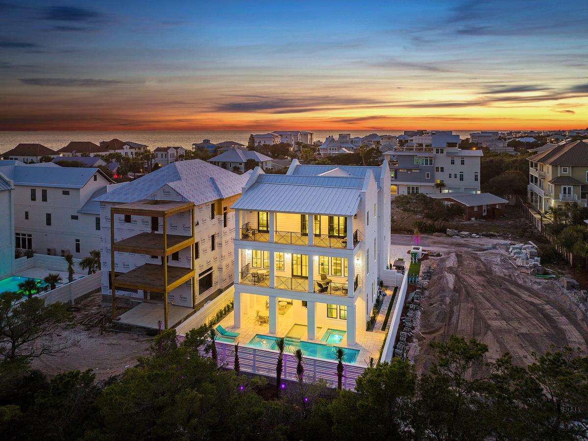 A modern, multi-story coastal home glows warmly at dusk, with a lit pool and spa in the backyard, surrounded by other beach houses and a view of the ocean on the horizon.