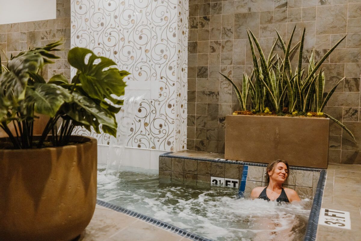 A woman relaxes in a warm spa pool as water flows gently from a tiled fountain wall. Soft lighting and lush plants make the space feel calm and inviting. It’s a quiet moment of island-style self-care.