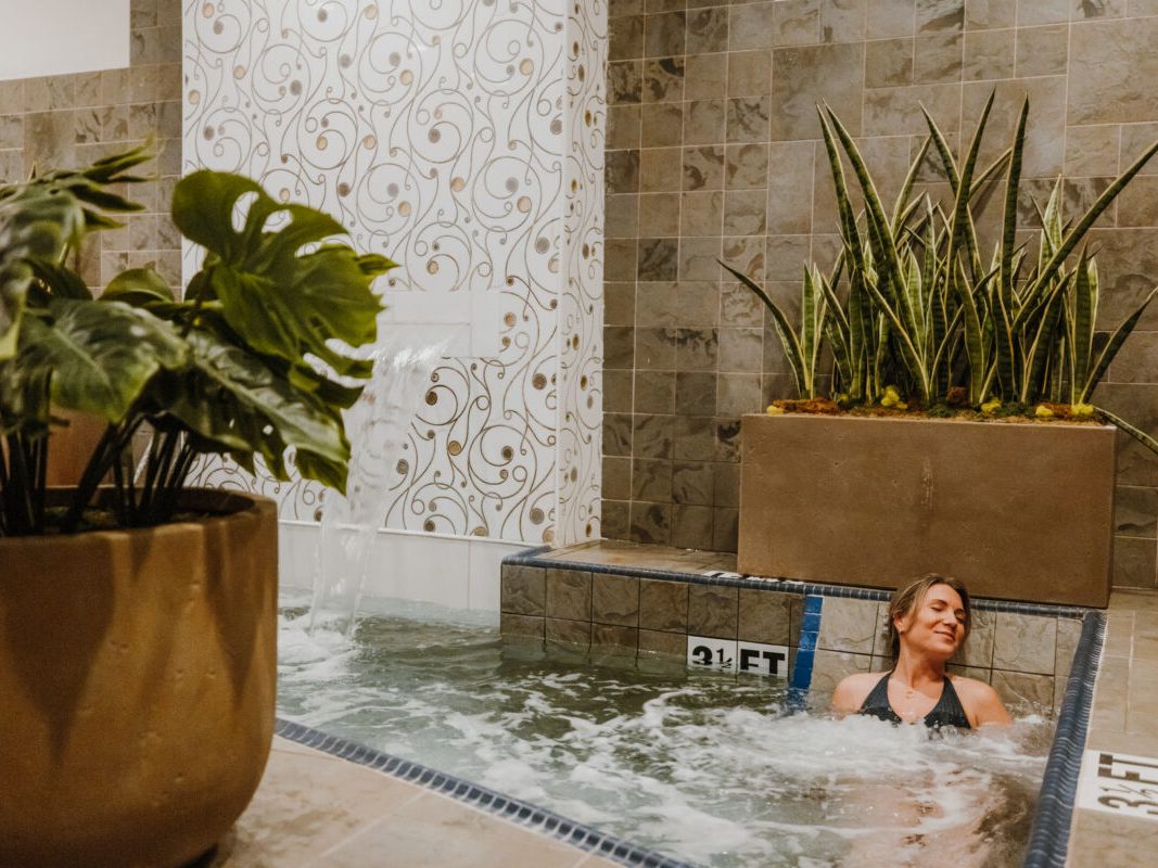 A woman relaxes in a warm spa pool as water flows gently from a tiled fountain wall. Soft lighting and lush plants make the space feel calm and inviting. It’s a quiet moment of island-style self-care.