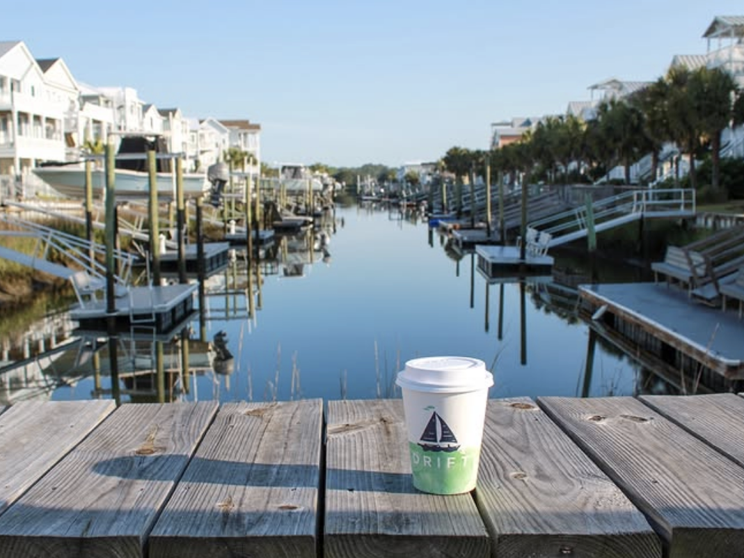 A to-go coffee cup from Drift Coffee sits on a wooden dock overlooking a peaceful canal lined with palm trees, boats, and coastal homes under a clear blue sky.
