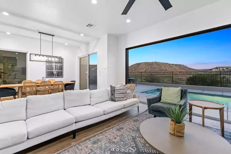 Bright living room with a white sectional sofa, modern dining area, and sliding glass doors opening to a pool and mountain views.