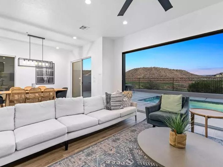 Bright living room with a white sectional sofa, modern dining area, and sliding glass doors opening to a pool and mountain views.