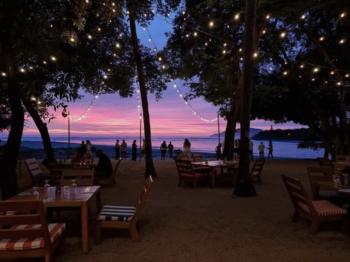 Beachfront restaurant with string lights hanging from trees, wooden tables on the sand, and a vibrant purple and pink sunset over the ocean