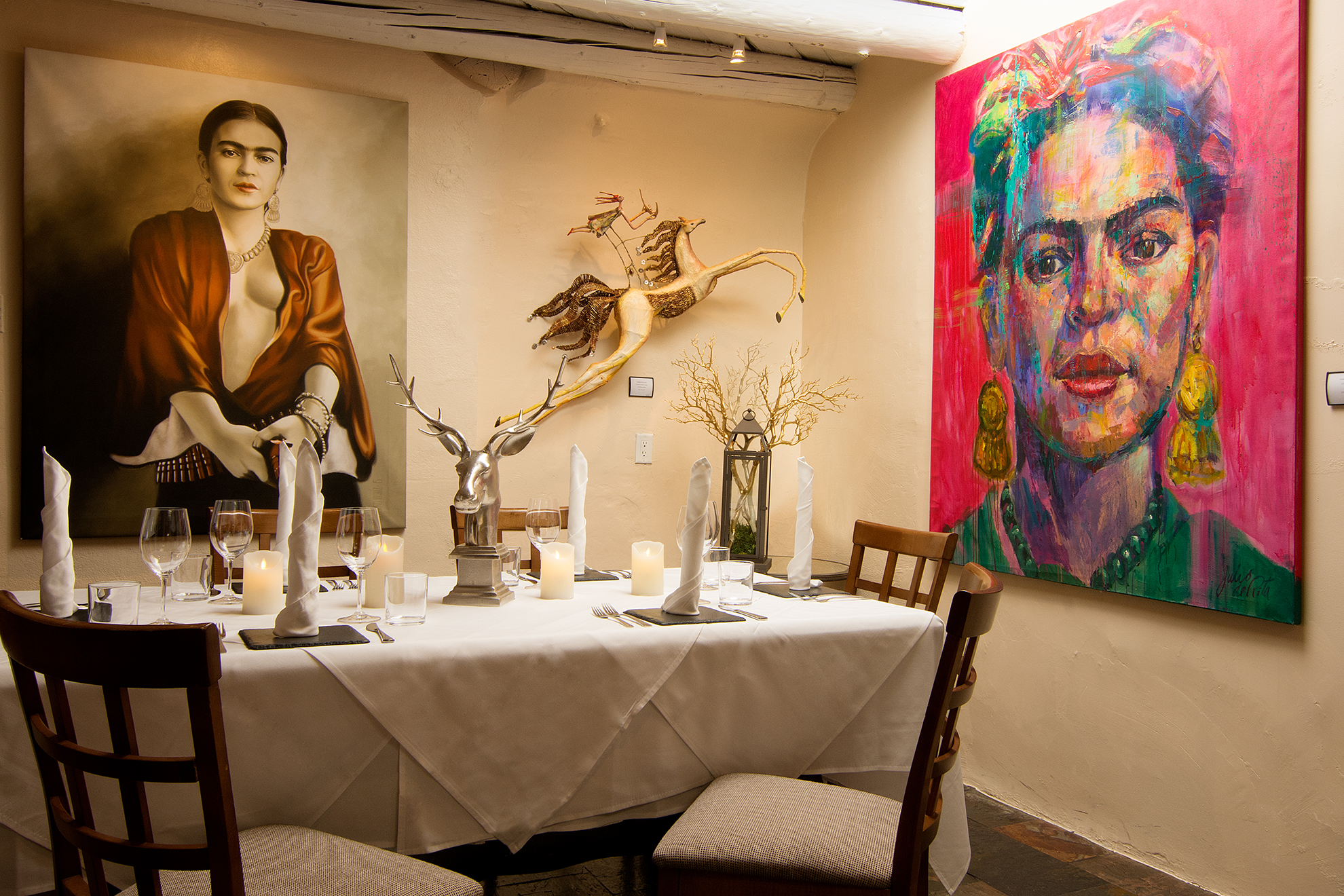 A cozy private dining room glows with soft lighting, surrounded by striking Frida Kahlo artwork. The white tablecloth, elegant place settings, and unique decor create an intimate, artistic atmosphere. It feels like a perfect spot for a memorable dinner in Santa Fe.