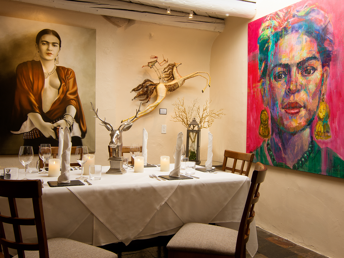 A cozy private dining room glows with soft lighting, surrounded by striking Frida Kahlo artwork. The white tablecloth, elegant place settings, and unique decor create an intimate, artistic atmosphere. It feels like a perfect spot for a memorable dinner in Santa Fe.