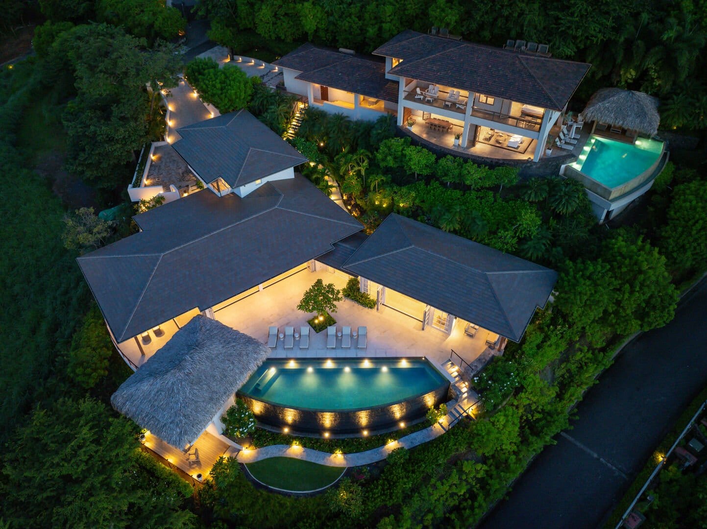 Aerial view of a luxury hillside estate at dusk featuring multiple villas, infinity pools, and lush tropical landscaping illuminated by warm outdoor lighting.