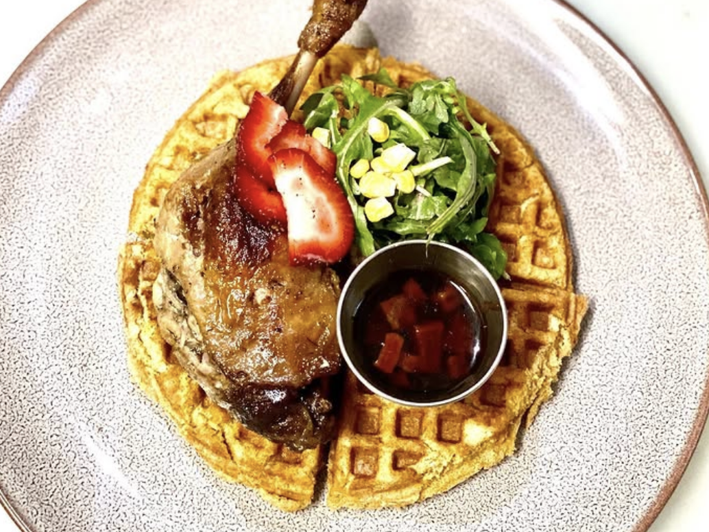 A gourmet twist on chicken and waffles featuring a crispy duck leg atop a golden waffle, garnished with fresh strawberry slices, a side of arugula and corn salad, and a small cup of syrup with diced fruit.