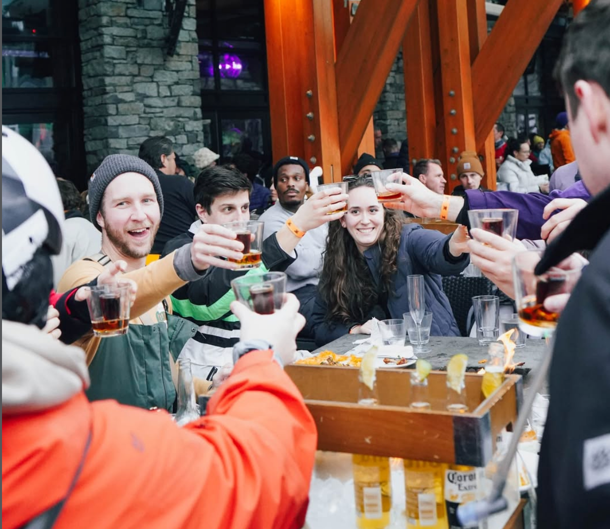 Group of friends gathered at an outdoor mountain bar, raising glasses in a celebratory toast amid a lively après-ski atmosphere.