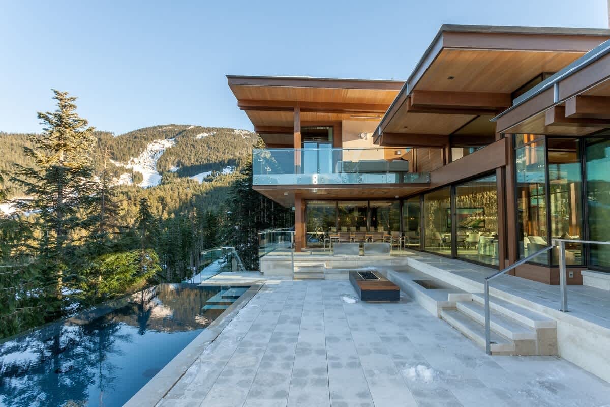 The striking exterior of Kadenwood Eagles Nest in Whistler features a contemporary architectural design with warm cedar ceilings, floor-to-ceiling glass walls, and a glass-railed upper balcony overlooking a sleek infinity pool and stone terrace. Snow-dusted mountain slopes and dense evergreen forest frame the luxury ski chalet against a clear winter sky.