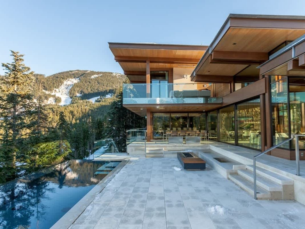 The striking exterior of Kadenwood Eagles Nest in Whistler features a contemporary architectural design with warm cedar ceilings, floor-to-ceiling glass walls, and a glass-railed upper balcony overlooking a sleek infinity pool and stone terrace. Snow-dusted mountain slopes and dense evergreen forest frame the luxury ski chalet against a clear winter sky.