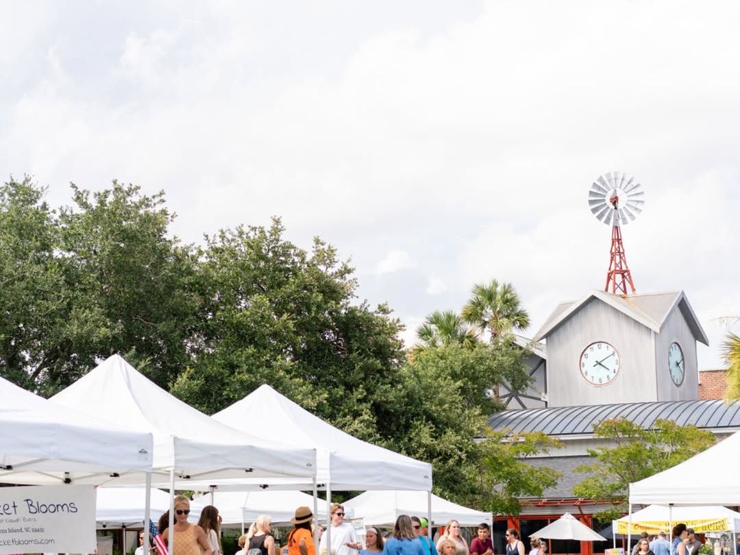 Shoppers browse through open-air market tents at Freshfields Village under sunny skies, with the iconic clock tower and red windmill in the background. A fun, local spot to explore on Kiawah Island.