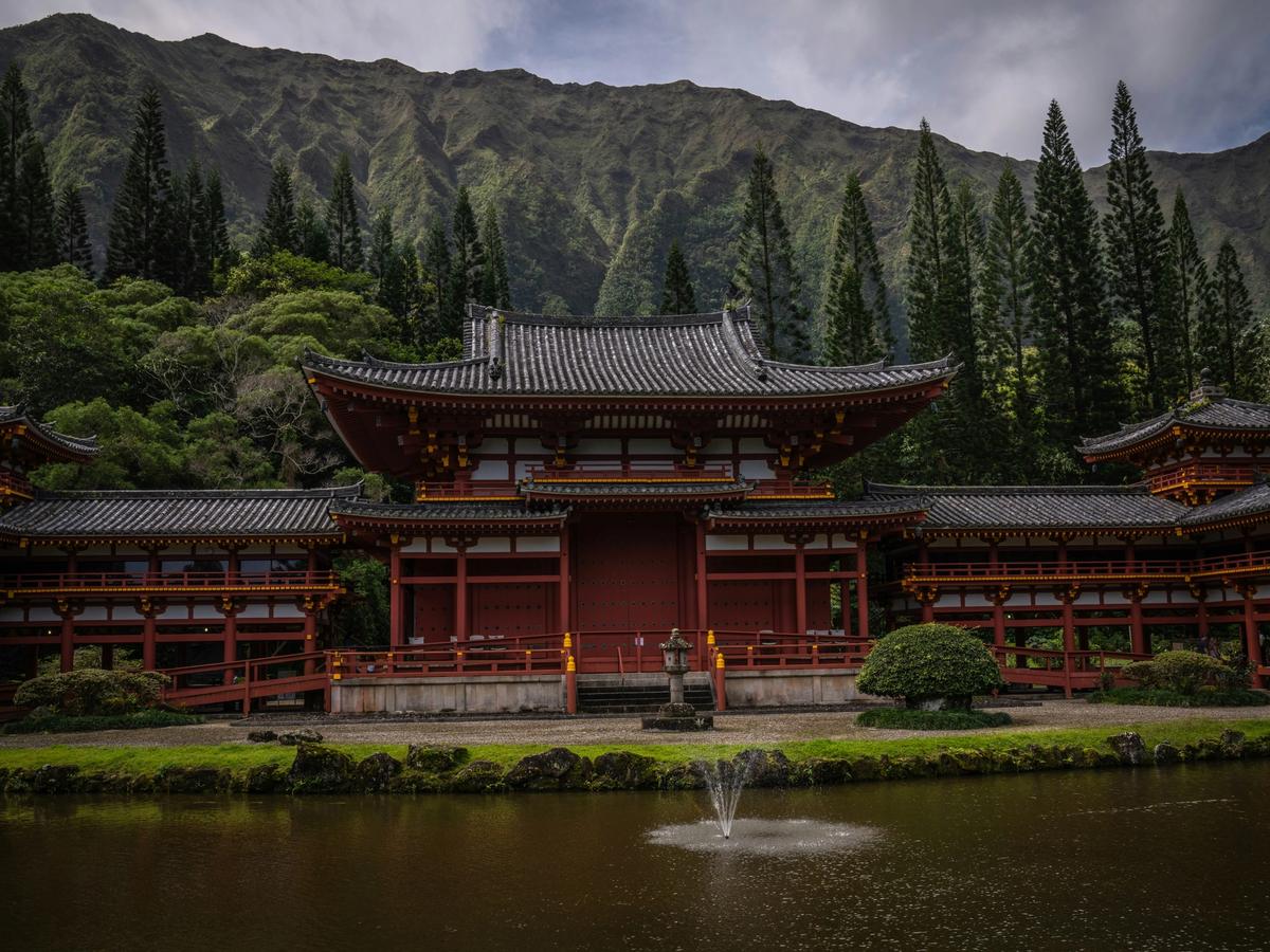 A traditional Japanese-style temple with ornate red and gold architecture sits peacefully beside a pond with a small fountain, framed by lush greenery and dramatic mountain ridges in the background.